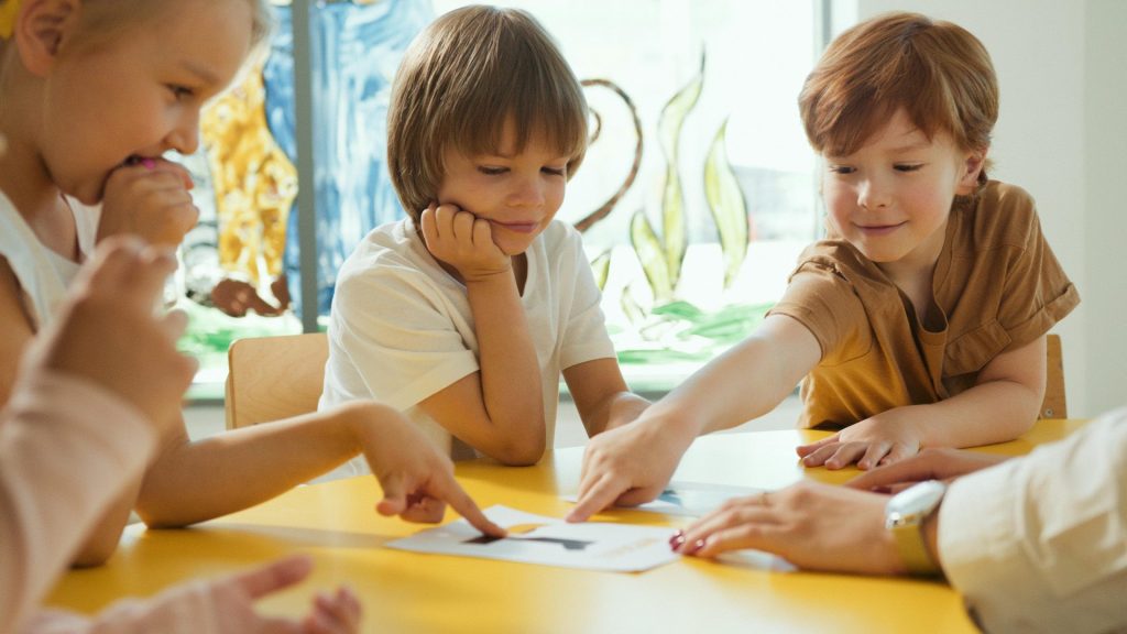 Four children and a tutor gathered around a table in an in-person group French class, smiling and interacting with an image, showcasing a fun and engaging learning environment.